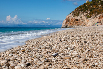 Fototapeta na wymiar Sunny blue Mylos pebble beach with azure sea, white foam and rocky cliff on coast of Lefkada island in Greece. Summer nature vacation travel to Ionian Sea