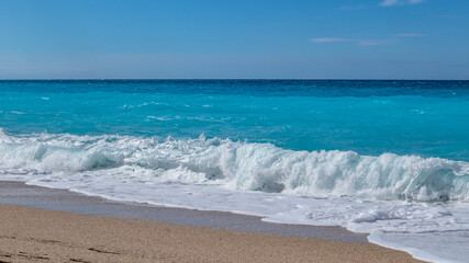 Fototapeta na wymiar Blue vibrant breaking waves on shore of Greek island with blue sky. Sandy beach in Greece. Summer nature travel to Ionian Sea