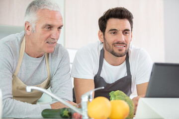 son and senior father in kitchen at home using tablet