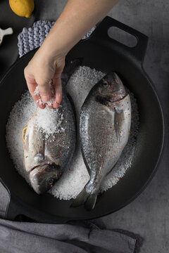 Woman Hand Dropping A Lot Of Coarse Salt Over A Pair Of Sea Breams In An Iron Cast Pan.