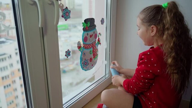 Child spray white artificial synthetic snow on window. Female person decorating living room to christmas and having fun indoors. Young girl kid prepare to new year holidays. Happy childhood concept