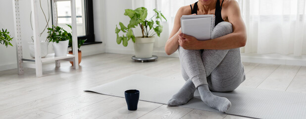 Young woman writing in her notebook. Sitting at home by the window on a yoga mat after exercise....