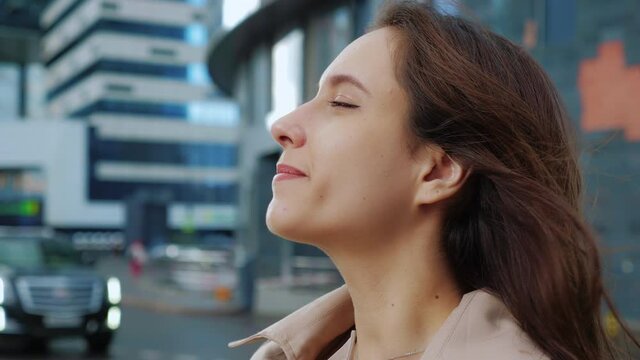 Portrait, profile, face of happy brown-haired lady is on street with office buildings. Joyful girl smiles, fools around, behaves like child, sticks out her tongue to taste raindrops. Autumn, rain
