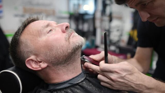Barber master shaving handsome mature bearded man using electric shaver in salon. Hair artist making beard style for person in male barbershop. Services of professional stylist. Fashion haircare for m