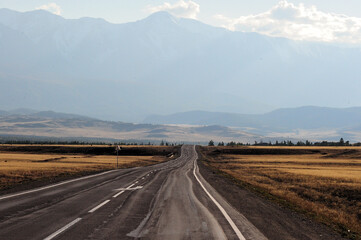 A winding road going through the autumn steppe to meet hills and snow-covered mountain peaks.