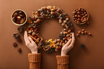 Female hands with beautiful acorn wreath and bowls of chestnuts and hazelnuts on brown background