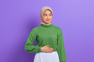 Beautiful smiling Asian woman in green sweater keep hands on the stomach and feel satiety after eating tasty isolated over purple background