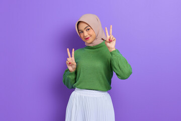 Obraz na płótnie Canvas Beautiful smiling Asian woman in a green sweater looking at camera and showing peace sign with fingers isolated over purple background