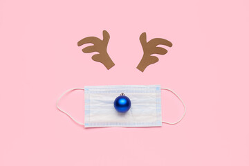 Composition made of toy reindeer horns, Christmas ball and medical mask on color background