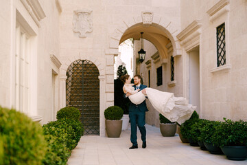 Groom carries bride in his arms in the courtyard of the house with arches