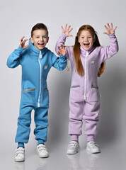 Two laughing, frolic kids boy and girl in blue and pink jumpsuits stand holding hands up, showing beast claws, playing, scare us over light background