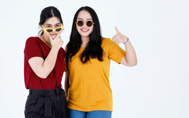 Studio shot of two young Asian cool fashionable stylish female friends in casual tshirt outfit wearing fashion vintage sunglasses standing smiling looking at each other eyes on white background