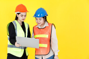 Studio shot of two Asian female professional engineering manager foreman wearing hard helmet and reflective safety vest holding paperwork document file folder discussing together on yellow background