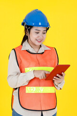Portrait studio shot Asian female professional engineering foreman manager in hard helmet and reflective safety vest smiling look at camera checking inventory stock from tablet on yellow background