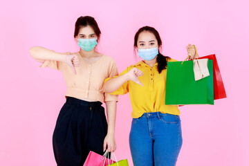 Portrait studio shot of two Asian young unhappy sad upset female shoppers friends wearing face mask and casual outfit carrying colorful shopping bags showing thumbs down together on pink background