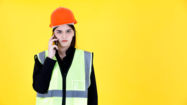 Portrait studio shot of Asian female professional engineering foreman manager with braces in hard helmet and reflective safety vest smiling talking on phone with colleague worker on yellow background - Powered by Adobe