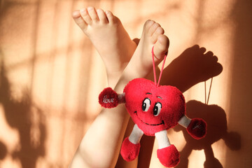 Female bare feet up with hanging heart toy on orange coral background with shadows lines