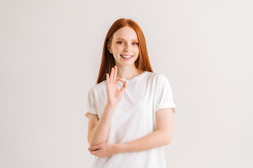 Portrait of smiling pretty redhead young woman showing okay gesture looking at camera, standing on white isolated background in studio. Fronv view of cheerful attractive female showing ok sign gesture