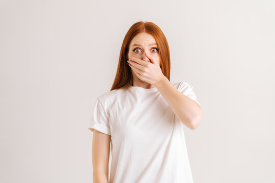Studio portrait of astonished shocked young woman covering mouth with pressed palms popping eyes at camera from surprise and worry, gasping witnessing terrible scene on white isolated background.