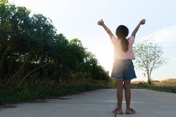 Little girl Thailand stood on the cement road with her back and shows arms up and looked at the evening sky in the background. with nature backgound of plantation cassava and grass.