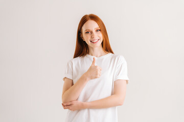 Fototapeta premium Portrait of cheerful pretty young woman showing thumbs up looking at camera, standing on white isolated background in studio. Front view of satisfied attractive female showing like gesture.