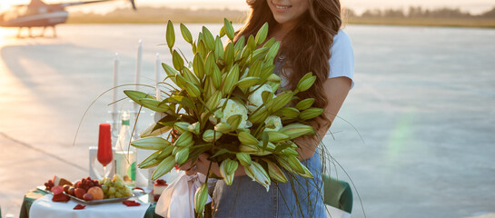 Woman holds a bouquet green lilies and white peonies in the background of table with fruits, wineglasses and candles. Girl with flowers. Proposal of marriage and small business concept. Copy space. © pijav4uk