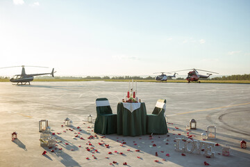 Table and chairs in green with fruits, glasses, wine and candles surrounded by white lanterns and rose petals with helicopters background. Proposal of marriage and small business concept. Copy space.
