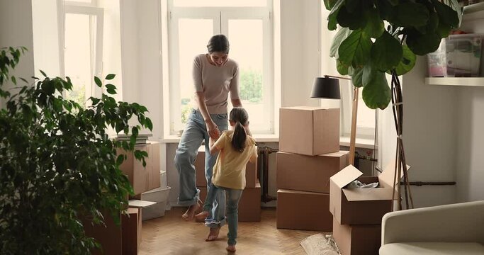 Happy laughing young single mom dance with preschool daughter at new home full of boxes with stuff celebrate relocating to modern apartment. Joyful mother little girl flat buyers tenants jump have fun