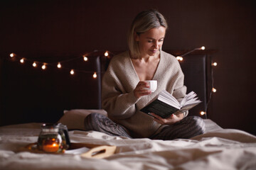 Adult attractive woman is reading book and drinking tea in her bedroom