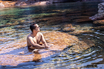 young man wearing sun glass sitting on rock in flowing river clear water at morning from flat angle