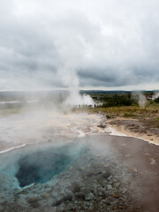 Steaming colorful hot spring pool with Strokkur geyser at the background in Geysir geothermal area, Iceland