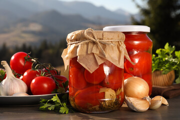 Glass jars with pickled tomatoes and fresh ingredients on wooden table against mountain landscape