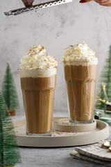 Grating chocolate over two tall glasses with cold coffee drink frappe - iced cappuccino with whipped cream, christmas decoration and trees