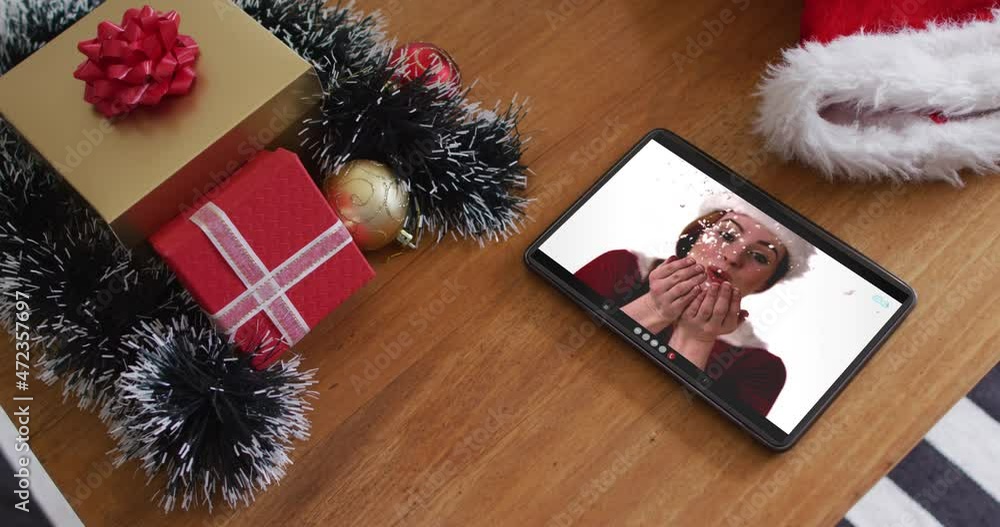 Poster Caucasian woman wearing santa hat and blowing snow on christmas video call on tablet