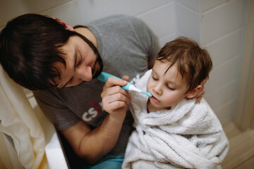 Father is brushing his little sons teeth. Baby boy is sitting on his father's laps wrapped up in a towel. Father's day.