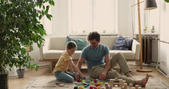 Young male babysitter sit on heated floor at living room entertain preschool girl assist in making fantasy constructions from diverse parts. Friendly dad and little daughter play colorful constructor