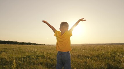 Fototapeta na wymiar happy child boy prays, spreading his hands to the sides at sunset, kid praying to the sun in sky, pulling helping hand, enjoying freedom outdoors walking in nature in park, inspiring childhood dream