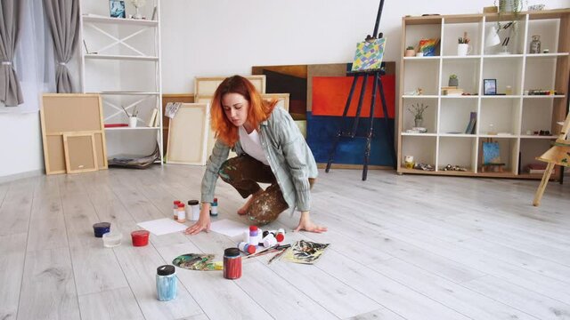 Art school. Female painter. Artistic tools. Serious woman preparing paper paint bottles and brushes sitting wooden floor in light room interior view.