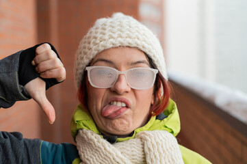 A disgruntled Caucasian woman stands on the balcony of a brick house in winter, showing her tongue and thumb down. Girl in ice-covered glasses on the street outdoors