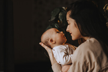 young brunette mom and her newborn baby in her arms, home photo in a dark room by the window