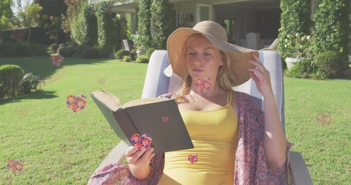 Animation of floral hearts falling over cauacsian woman in sun lounger reading book in garden