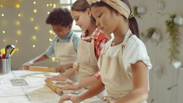 Tilt Up Shot Of Little Asian Girl In Apron Rolling Dough On Kitchen Table And Playing With Rolling Pin During Kids Cooking Class