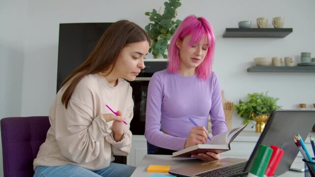 Lovely young woman with birth hands anomaly and attractive pink haired female friend preparing for university exams, studying and discussing educational material together in college dorm room.