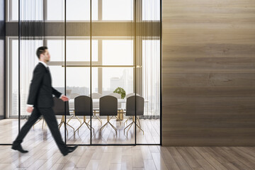 Businessman walking in modern wooden meeting room office interior with empty mock up place on wall, table, armchairs, window with city view and daylight. Workplace and law and legal concept.