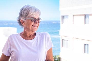 Portrait of a mature attractive gray-haired woman relaxing in her home balcony, background of the sea and blue sky during a sunny day outdoors.