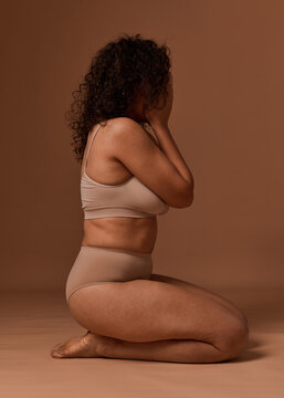 Depressed woman in lingerie with cellulite and stretch marks closes her eyes, sitting against a beige background, does not accept herself and her body because of the framework established by society.