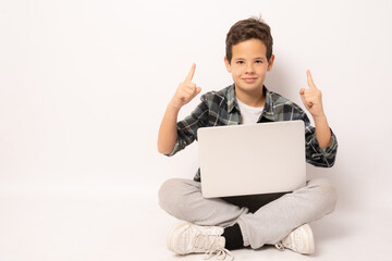 Smiling cute boy wearing plaid shirt sitting on floor holding laptop computer pointing fingers up isolated over white background.