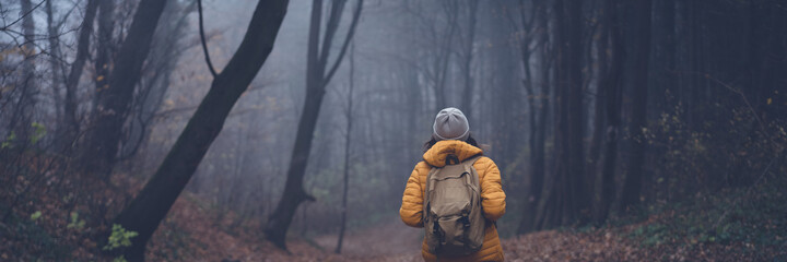 Young woman traveler in a yellow jacket with backpack standing in the foggy forest with her back. Banner format