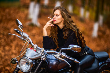 Fototapeta na wymiar A beautiful long-haired woman smoking on a chopper motorcycle in autumn landscape on a forest road