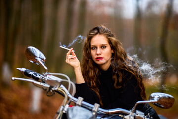 A beautiful long-haired woman smoking on a chopper motorcycle in autumn landscape on a forest road